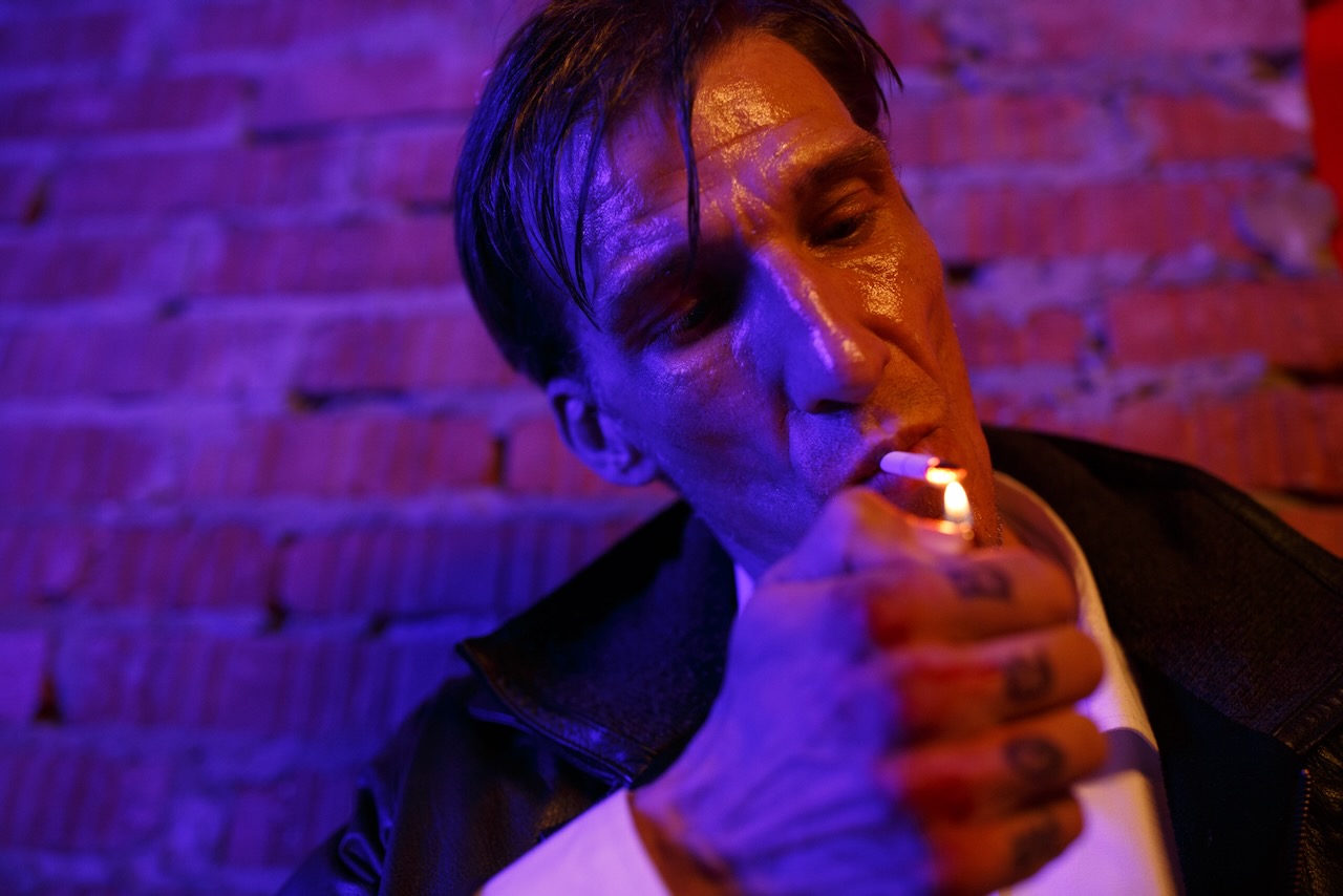 A man lighting a cigarette against a brick wall, lit in blue and red.